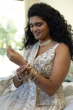 Indian woman sitting by window on bed adjusting gold bangles while wearing embroidered dress