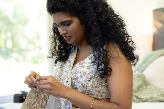 Woman fastening delicate gold necklace at dressing area while wearing pale sheer drape and bangles