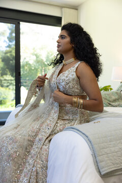 Indian woman sitting on bed in bedroom, wearing embroidered lehenga and adjusting sheer dupatta