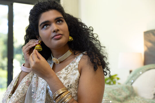 Indian woman adjusting gold jhumka earrings and wearing embroidered saree in bedroom