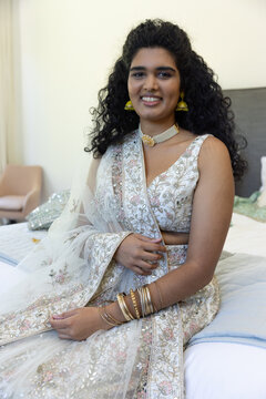 Adult Indian woman sitting on bed holding sheer dupatta, wearing pastel lehenga and gold jewelry