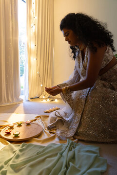 Indian woman kneeling and placing lit diyas on round tray by curtained glass door, in lehenga