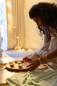 Indian woman kneeling arranging lit diyas on tray by lights wearing embroidered ivory bangles