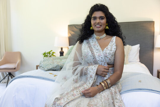 Indian woman sitting on bed in room fixing dupatta while wearing lehenga,choker,jhumka,bangles