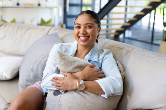 African American woman sitting on sofa at home wearing denim jacket wristwatch hugging pillow