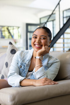 Woman smiling, resting hand on cheek, sitting on beige sofa wearing denim jacket near glass doors