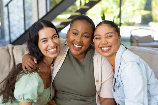 Diverse female friends laughing and hugging on beige couch in bright modern living room