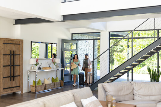Adult female friends entering home entryway carrying rolling suitcases, holding hat and tote bag