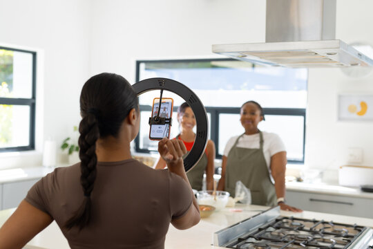 Diverse female friends filming at kitchen island using ring light and smartphone, wearing aprons