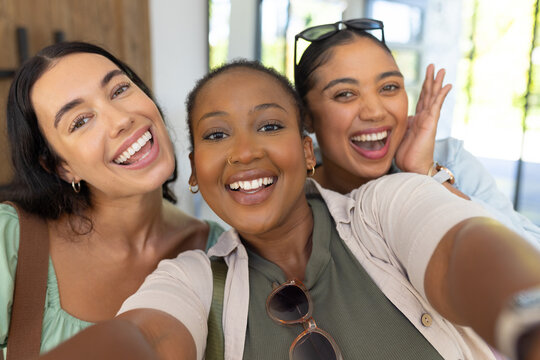 Diverse female friends posing, smiling and holding smartphone with sunglasses in bright lobby