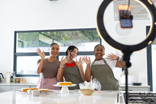 Female friends waving, decorating cakes and streaming with ring light smartphone in modern kitchen