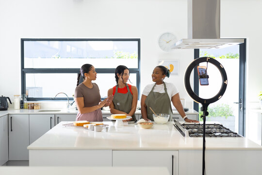 Diverse female friends filming while preparing cake in home kitchen on white island with ring light