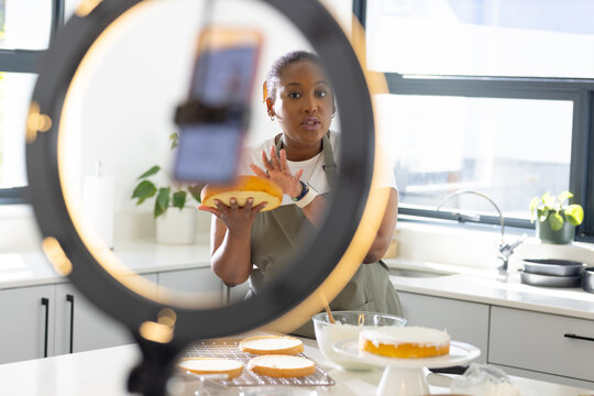 Woman holding cake layer, gesturing while recording with ring light and smartphone in kitchen