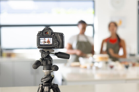 Female cohosts in aprons filming and preparing cake in kitchen-studio with DSLR camera on tripod