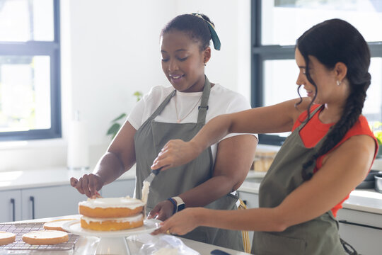 Diverse female friends frosting two-layer sponge cake in bright kitchen with offset spatula, aprons