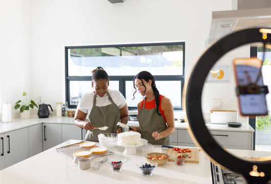 Diverse female friends wearing aprons frosting layered cake on white kitchen island with ring light