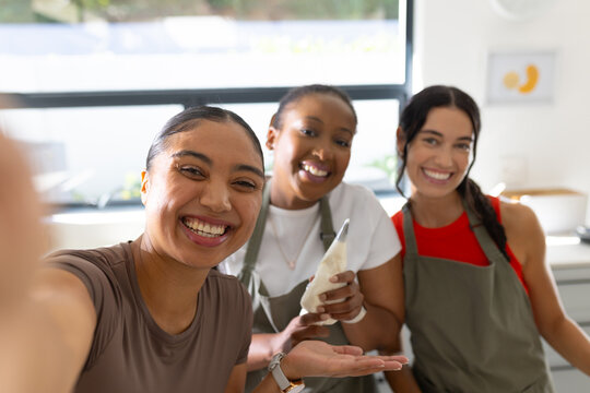 Diverse female friends taking selfie in kitchen wearing aprons with smartphone and piping bag