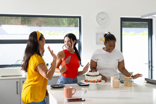 Diverse female friends laughing and cutting berry cake on white kitchen island holding metal server
