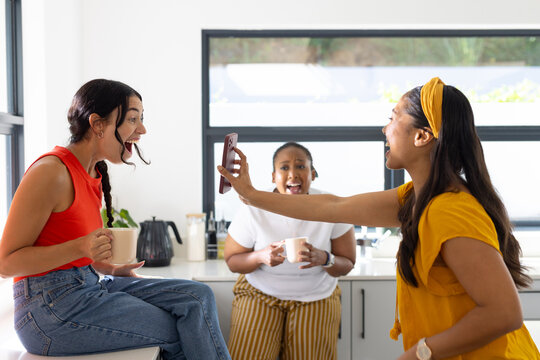 Diverse female friends laughing, pointing smartphone and holding ceramic mugs in modern kitchen