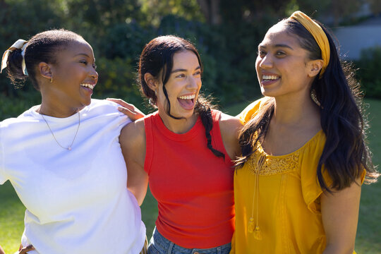 Diverse female friends laughing, linking arms on lawn, showing pendant, red top, mustard headband