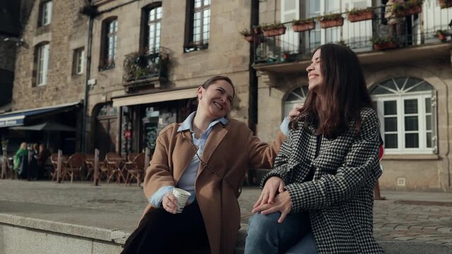 Two young women enjoy a cheerful afternoon together in a picturesque European town. Smiling, laughing, and animatedly chatting, they share authentic emotions and a joyful friendship in a relaxed outdo