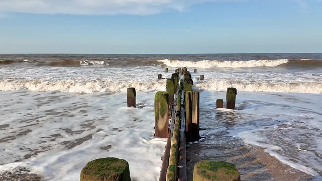 Pull back shot of waves rolling onto the shore, weaving around weathered wooden groynes as sea foam drifts gently across the beach. Slow motion.