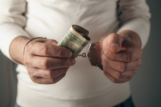 A man's hand holding a criminal's handcuffs, the concept of legality, and holding a dollar as a bribe