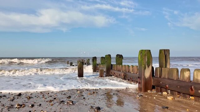 Pull back shot of waves rolling onto the shore, weaving around weathered wooden groynes as sea foam drifts gently across the beach. Slow motion.