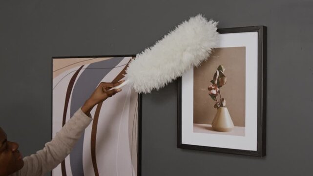 African American girl using white feather duster to clean two framed artworks hanging on dark grey wall