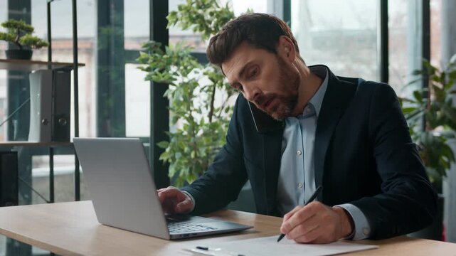 Busy successful Caucasian businessman typing laptop talking mobile phone office manager nervous male multitasking business man guy communicate client writing notes explaining using cellphone indoors