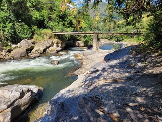 Fototapeta premium Bridge over the Macaé River rapids, Encontro dos Rios, Lumiar, Nova Friburgo, Serra Fluminense, Brazil; Atlantic Forest, clear waters, lush foliage, south american rainforest, adventure destination.