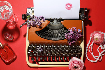 Fototapeta premium Vintage typewriter with paper, flowers, jewellery, perfume and glass of water on red background, flat lay