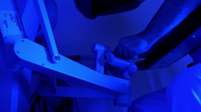 Male hands move the manipulators of the robotic surgical system. Unrecognized medic works in the room with blue light.