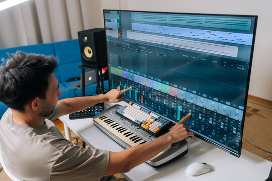 Rear view of sound engineer male creating new music in home studio, arranging tracks and mixing audio levels with midi keyboard and software on a large display, showcasing modern music production.