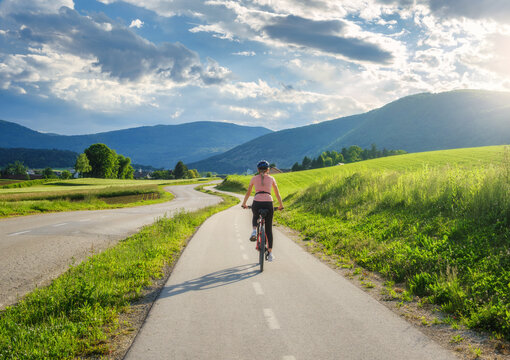 Woman riding mountain bike on asphalt bicycle path in Slovenia countryside in summer. Female cyclist cycling on lane through green meadows and hills at sunset. Summer outdoor activity, fitness. Travel