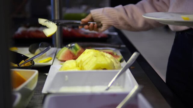 Woman's hand using tongs to pick up a slice of fresh melon from a self service buffet in a hotel restaurant, choosing healthy food for a balanced continental breakfast on vacation
