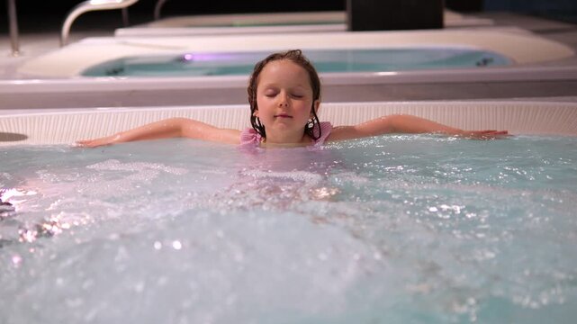 Calm young girl enjoying warm bubbling water while floating on her back in a spa pool, creating a serene and peaceful atmosphere of childhood relaxation and wellness