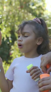 Vertical video: Checking wand, girl in pink blowing bubbles in yard as adult dipping orange bottle