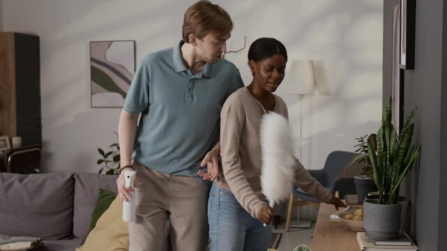 Multiethnic young couple doing housework, dancing energetically and singing, man holding spray bottle like microphone, woman with feather duster