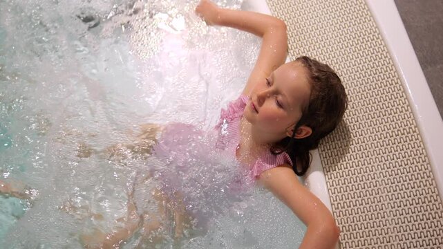 Young girl lying back and relaxing in bubbling spa water, enjoying warm jets and soothing foam as she calmly rests at the pool edge during a playful and peaceful wellness moment