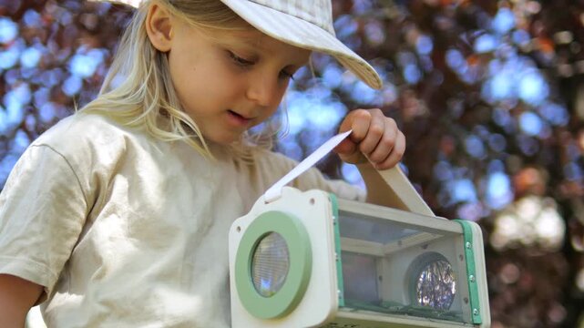 Young explorer with long blonde hair and a cap intently studying insects inside a wooden bug viewer box, fostering a love for nature and science education through outdoor play and discovery