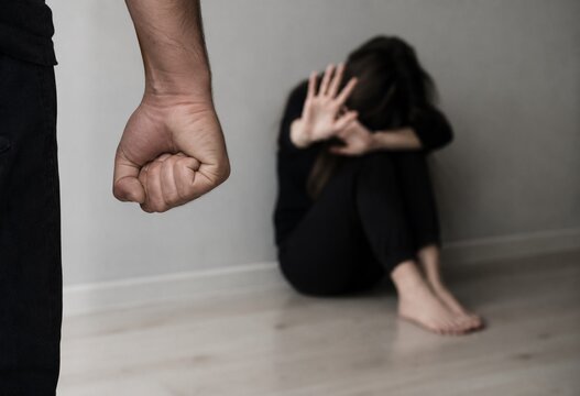 Conceptual image of domestic abuse showing a man's clenched fist in sharp focus in the foreground, with a terrified woman cowering in the background corner.