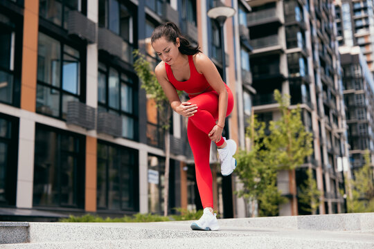 Attractive young woman feeling pain in her foot during sport workout outdoors. Girl in red sportswear and white sneakers standing on one leg and feeling cramps in her lef leg. Urban sport concept.