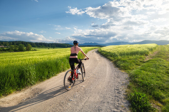 Woman riding mountain bike on dirt road through green wheat fields at sunset in Slovenia. Female cyclist cycling in countryside landscape and sunny sky. Summer outdoor activity, fitness. Girl on bike