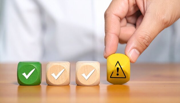 Hand Placing Yellow Warning Attention Block Next to Row of Green Check Mark Cubes Representing Risk Management Quality Control Assessment and Evaluation Concept