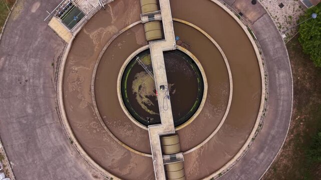 Top-down view of a circular secondary clarifier surrounded by concentric aeration basins at a municipal sewage treatment facility, showing brown effluent and radial walkways