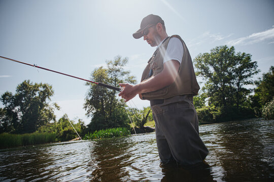 Young fisherman fishing on lake or river. Adult guy in fisherman's clothes and cap holding rod in hands. Using lure for catching fish. Mand stands in river or lake water. Beautiful sunny day.