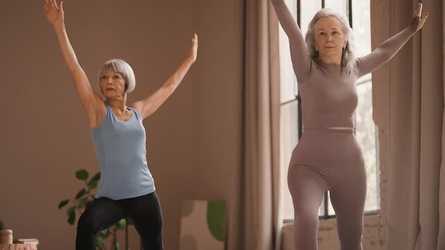 Two senior female friends practicing yoga together, performing warrior pose