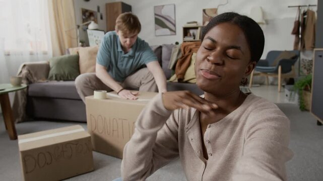 POV of cheerful African American gen Z girl speaking on camera while recording video with Caucasian boyfriend sealing donation box in background