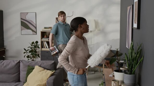 Black girl using cleaning duster and speaking with Caucasian boyfriend vacuuming rug while couple doing housework together